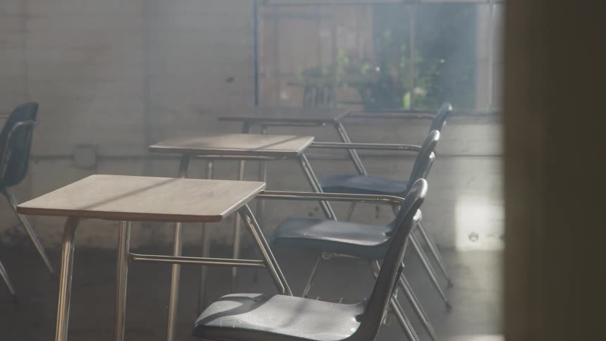 Empty desks and chairs sit in a sunlit classroom with a soft haze and exposed brick walls