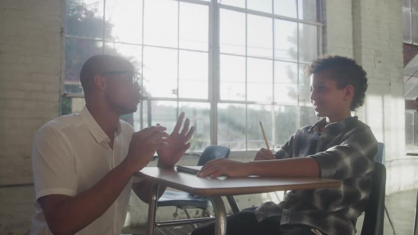 Man explaining to boy sitting at school desk in room lit by large window