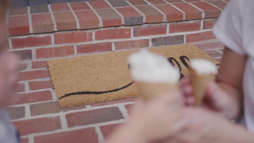 Children exchange ice cream cones and eat them on a doorstep with a doormat that reads hello