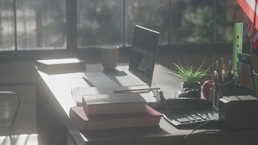 An office desk equipped with a laptop books an american flag and various office supplies is bathed in sunlight from a nearby glass window with trees visible outside covering the road and a car driving by at midday