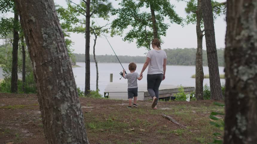 A woman holding her son walks toward a dock through the woods with the young boy holding a fishing rod at midday