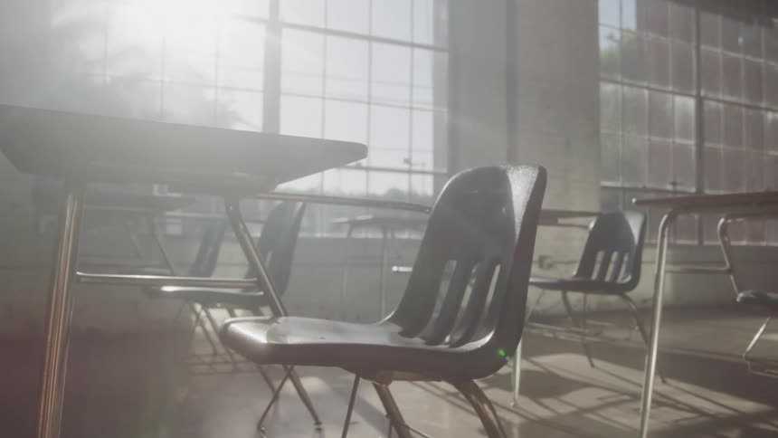 The video shows an empty classroom with a hazy ethereal quality a single chair is prominently featured in the foreground with several other desks and chairs visible in the background all bathed in bright sunlight streaming through large windows