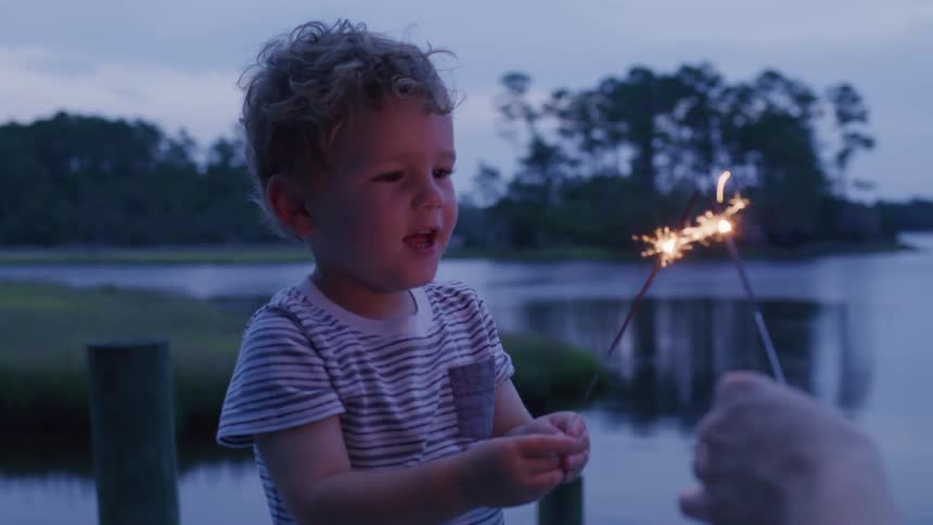 A closeup of a happy young child on a dock at dusk looking at and holding lit sparklers while another person assists