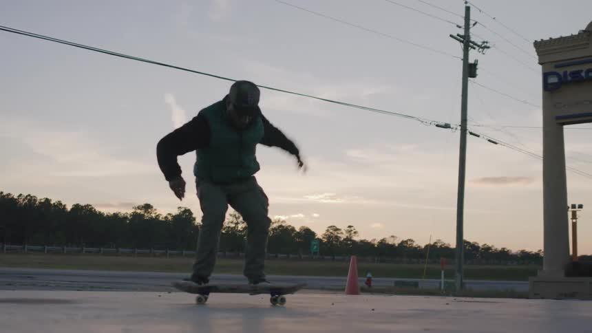 A person wearing a dark vest and grey pants performs a trick flipping the skateboard beneath them while standing on a paved surface