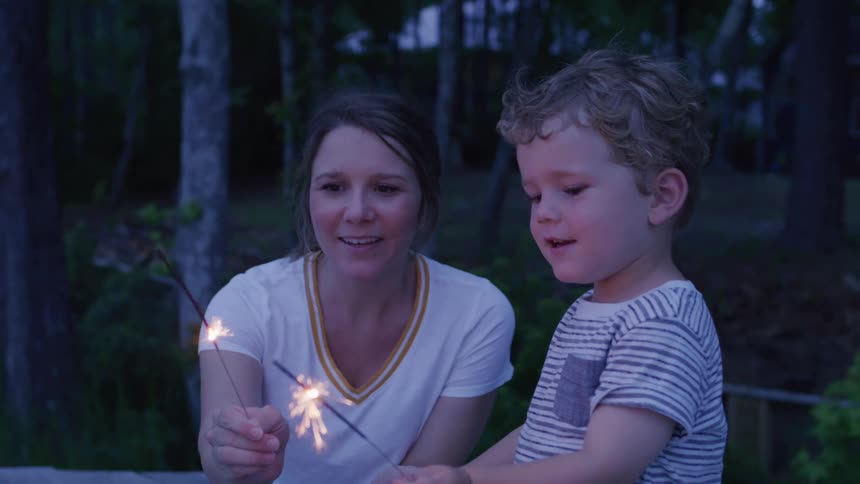 A woman and a small boy smile as they hold lit sparklers in front of them with a dark background of trees and a building