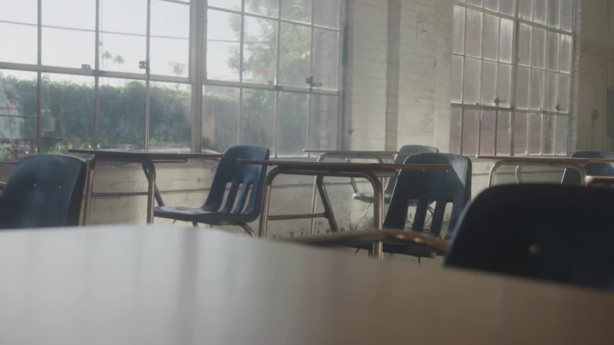 A view from the front of a classroom shows several rows of empty desks and chairs next to large windows