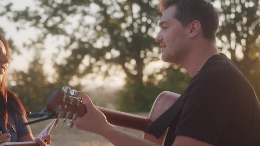 A young man sits in a grassland with a young woman playing a guitar at sunset both having fun as the man sings