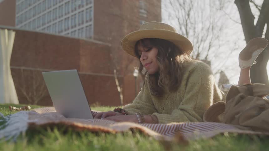 A young woman is lying on a blanket on a grassy lawn with a laptop in front of her she is wearing a hat and a fuzzy sweater and an office building is visible in the background