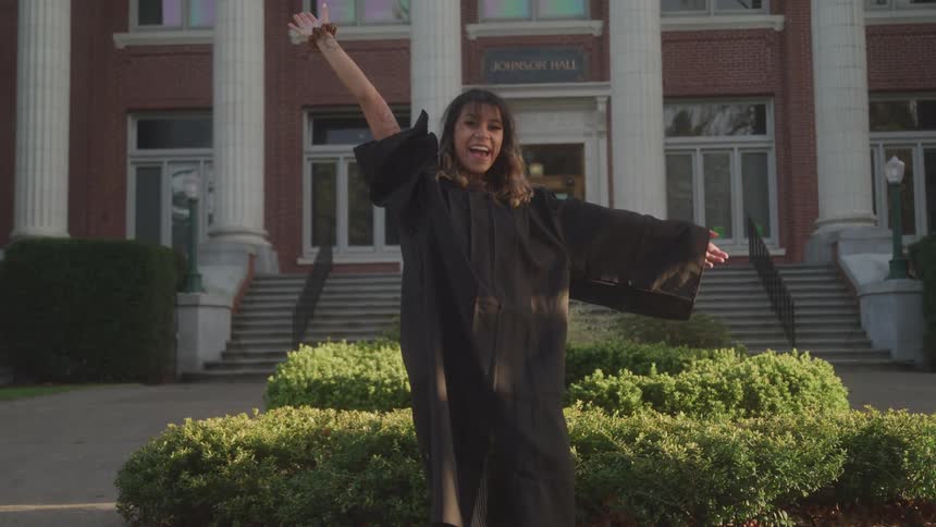 A woman in a black graduation gown stands outdoors on a sunny day in front of a large brick building and excitedly waves her arms
