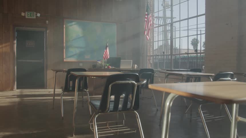 Sunlight streams through tall windows into an empty classroom with wooden walls black chairs and american flags