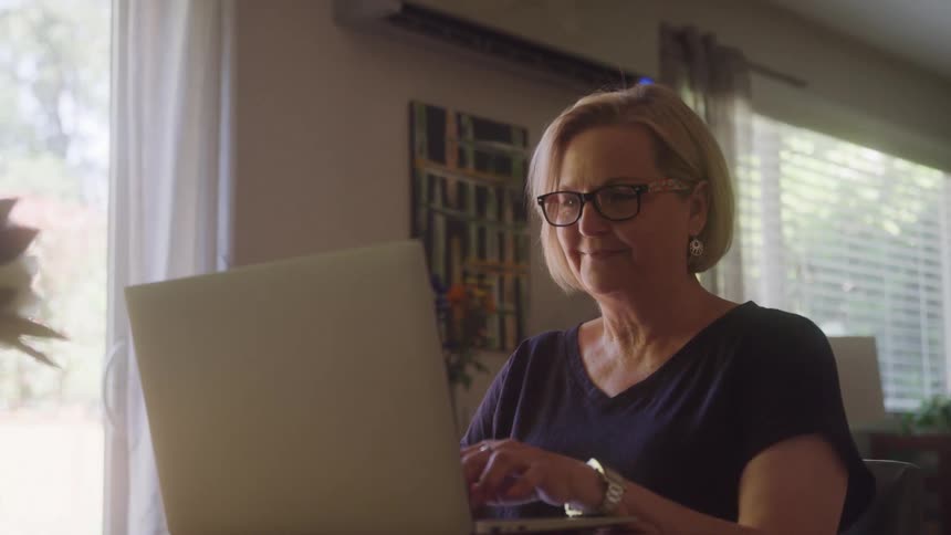 Blonde woman wearing glasses types on a laptop while sitting near a window lit by daylight