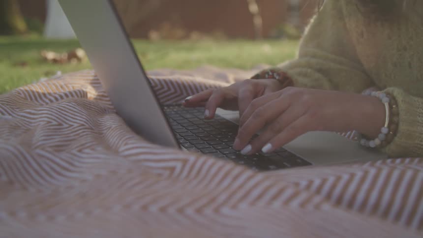 A woman lays on a blanket in her yard while using a laptop