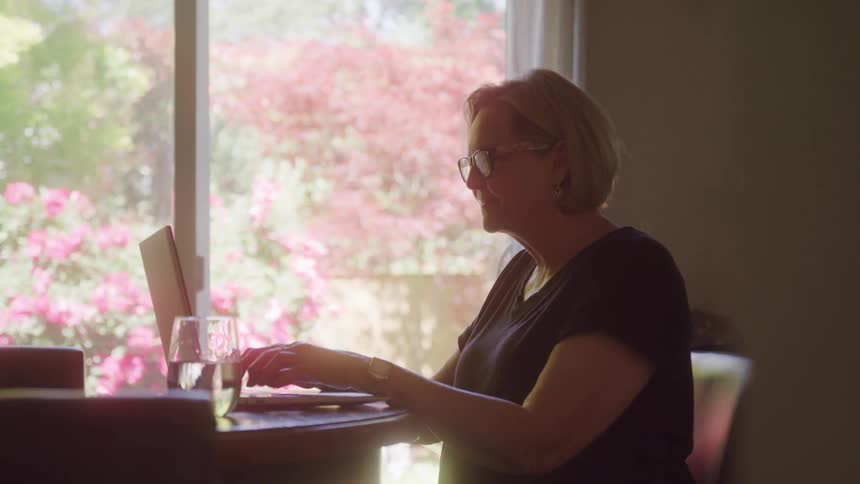 Woman with glasses types on a laptop near a window with flowers outside