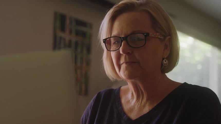 A woman in glasses looks at a laptop thoughtfully in warm indoor lighting