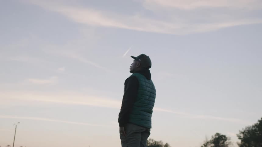 A man in outdoor gear gazes upward at a pastel evening sky with a jets contrail above
