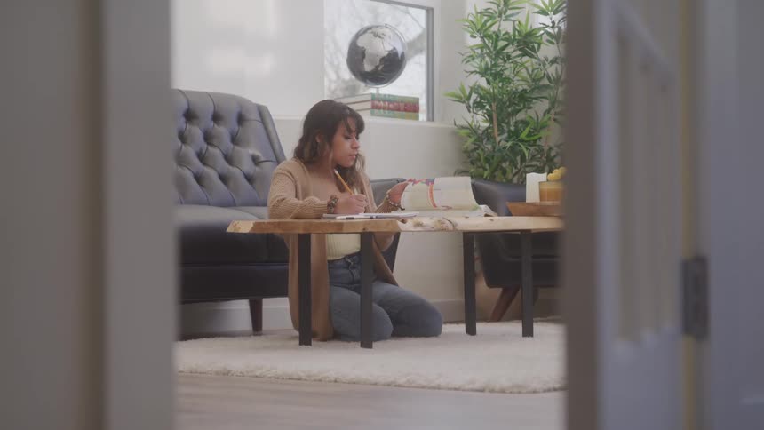Young woman kneeling on the rug writes in a notebook on the wooden table