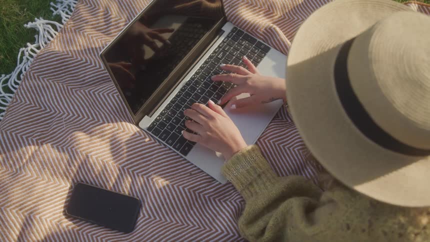 A woman wearing a hat and a sweatshirt sits on a blanket on a lawn typing on her laptop keyboard under the sun