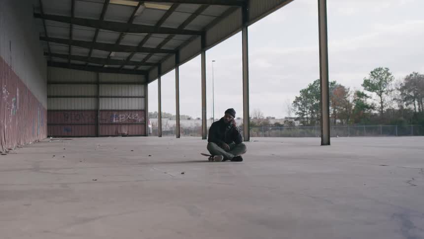 A man sits on the floor under an outdoor warehouse making a phone call at midday