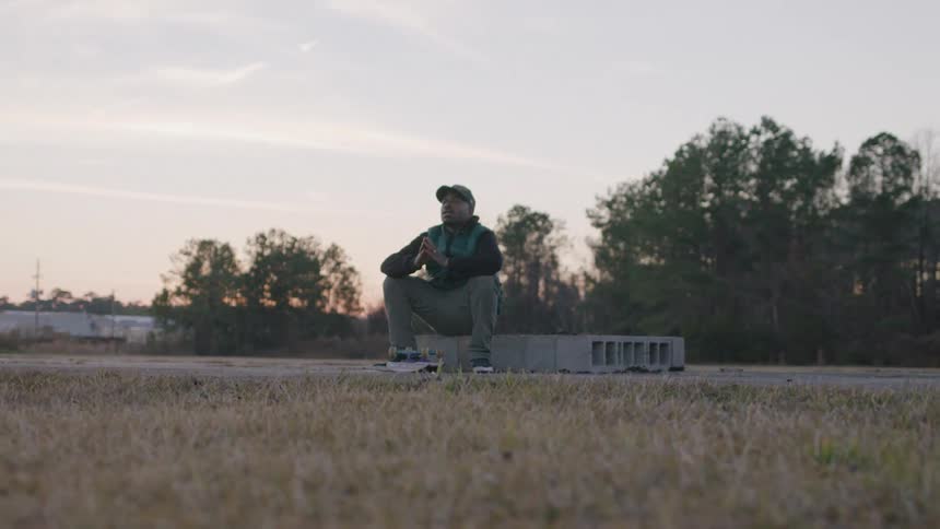 A man wearing a dark jacket a baseball cap and a green backpack sits on a concrete block in a grassy field