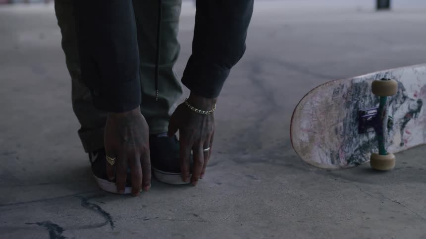 A man with tattoos and jewelry stretches near his skateboard on a concrete surface