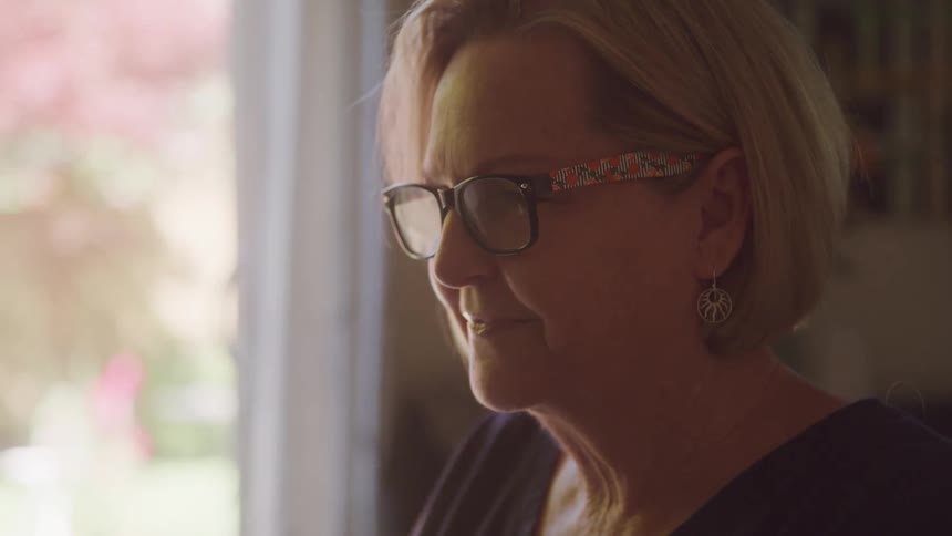 An elderly white woman sits indoors wearing glasses with a smile on her face at midday