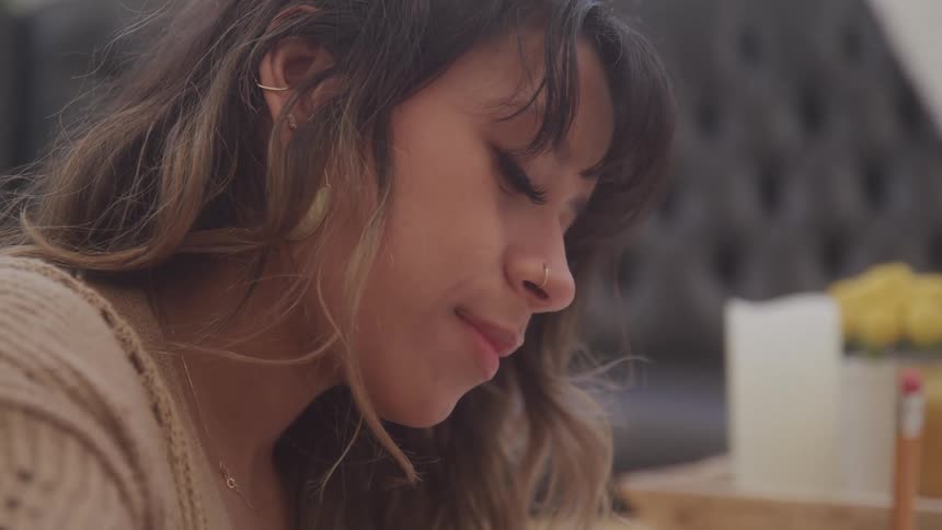 A woman with long wavy brown hair and highlights looks down while writing with a pencil with a focused expression