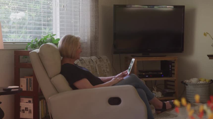 A woman relaxes in a comfortable white chair in a sunlit living room scrolling through content on a smartphone