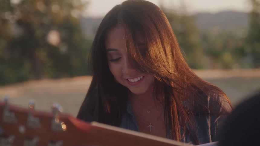 A woman with long dark hair smiles while looking down reading while a man plays guitar next to her