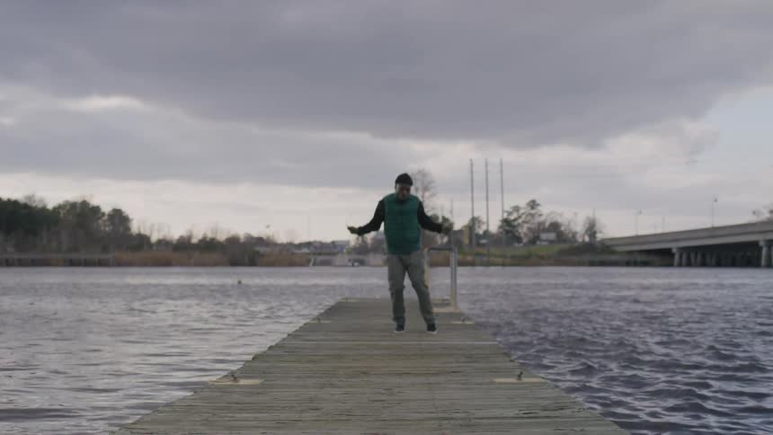 A man in a dark vest and beanie jumps rope on a dock under a cloudy sky by the water