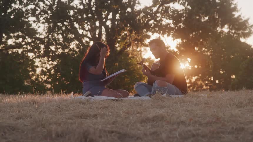 A couple sits in a grass field as the man sings and plays guitar while the woman writes on a notepad both enjoying their time together at sunset