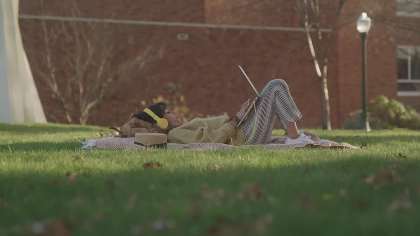A young woman lies on a blanket on the lawn wearing yellow headphones typing on her laptop at midday