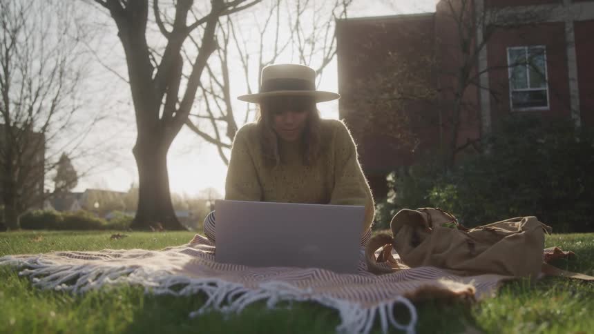A person wearing a wide brimmed hat sits on a blanket on a grassy lawn with a laptop in front of them working in the bright sun