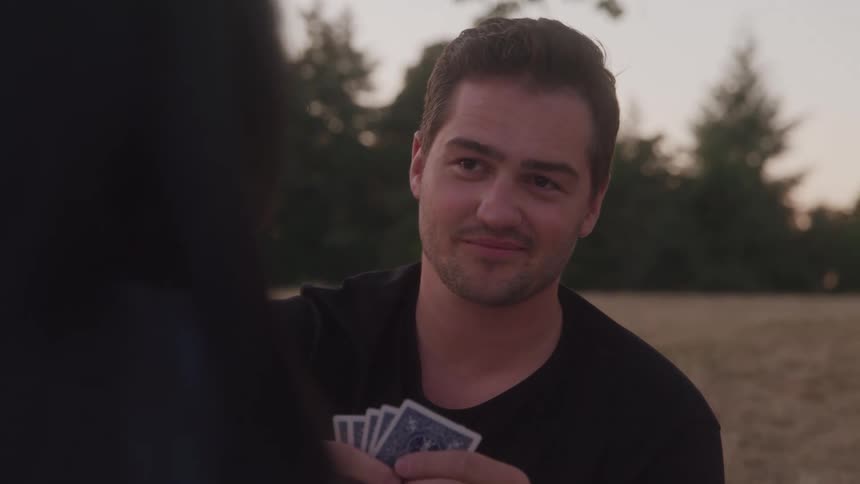 A man and woman are playing cards outside the man picks up a card looks at his hand and smiles and says something to the woman