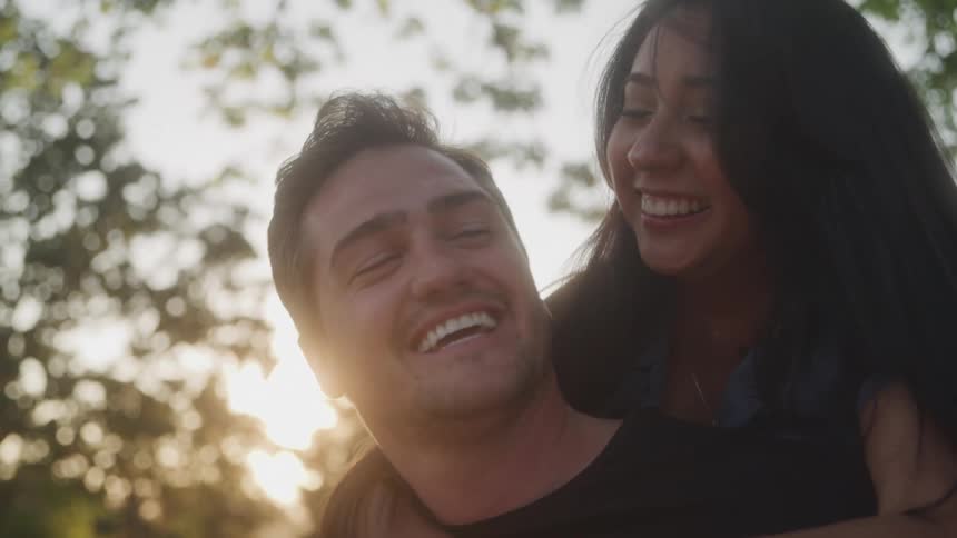 A woman with long dark hair is riding on a mans back and they are both smiling and laughing in an outdoor setting the background is out of focus with blurry sunlight filtering through trees