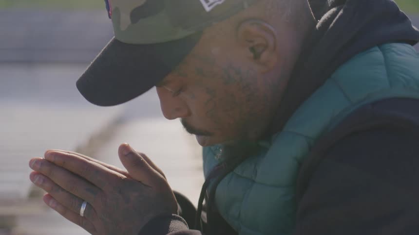 Man in a cap holds his hands together in a prayer gesture outdoors