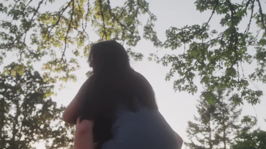 A woman with long dark hair smiles while getting a piggyback ride from a man with short brown hair as they are framed by sunlit trees