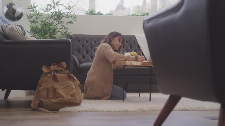 A woman wearing a tan cardigan kneels on a white rug in a sunlit living room writing on a notepad while a backpack rests on the floor next to her