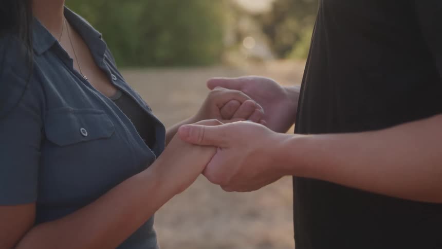 A couple stands on a grassland holding hands and facing each other at midday
