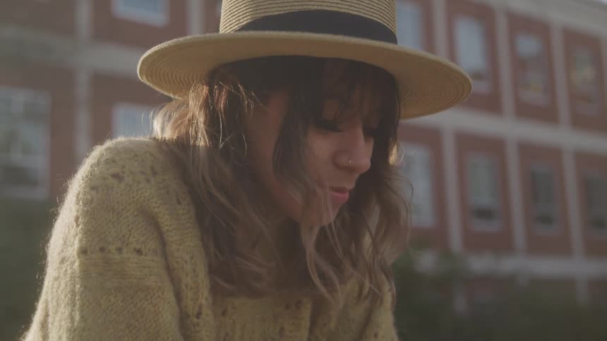 A young woman wearing a sweatshirt and a hat stands on a yard in front of a building