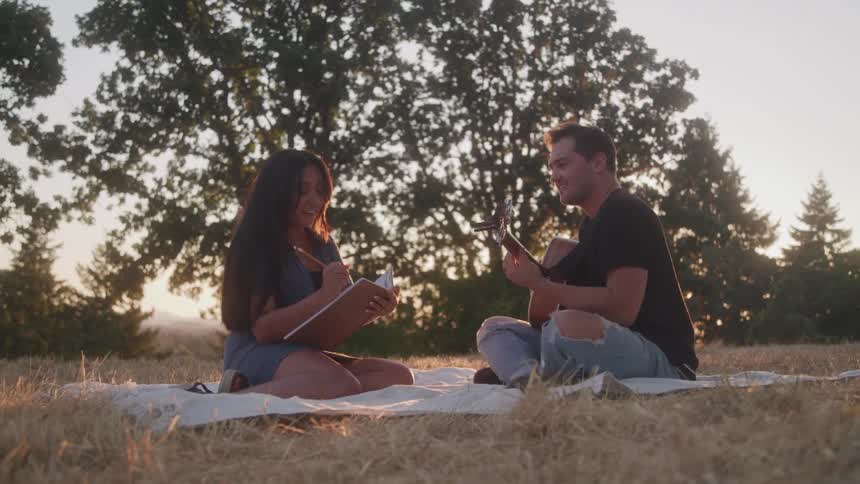 The video shows a couple on a picnic blanket in a field with trees in the background at sunset the man is playing a ukulele while the woman is writing in a notebook and they are both smiling and enjoying each others company