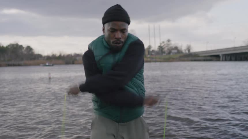 A man in a black beanie and a teal vest is actively working out with a jump rope by a body of water under a cloudy sky