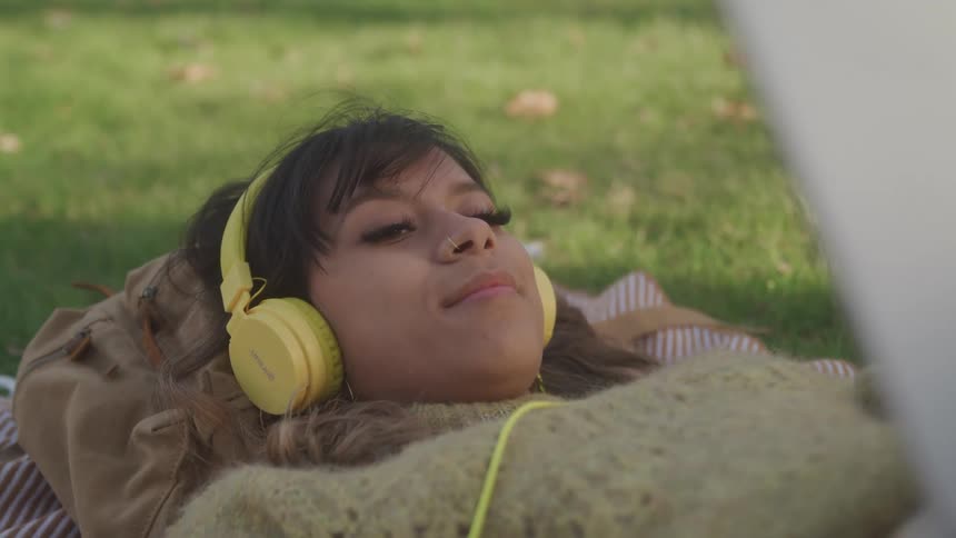 A woman wearing yellow headphones is lying on a blanket on the grass appearing to be relaxing or listening intently to music