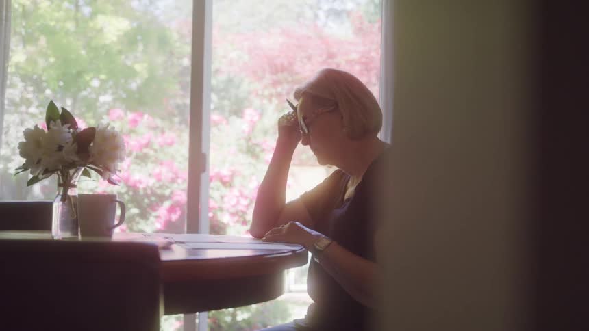 An elderly woman sitting at a table one hand holding a pen resting against her head and the other hand on a sheet of paper as she reads it