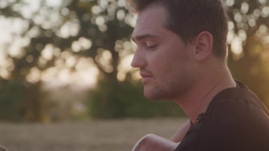A young man sits on a grassland holding and playing a guitar while singing at sunset