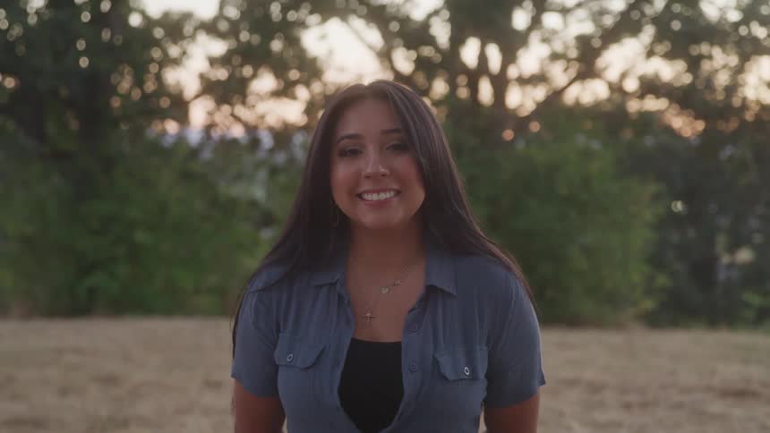 A smiling young woman stands outdoors at sunset wearing a blue shirt and layered necklaces