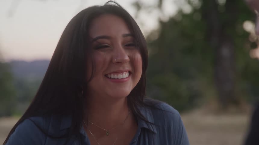 A woman in a blue top is smiling and probably laughing in a park someone is with her but we only catch a glimpse of them
