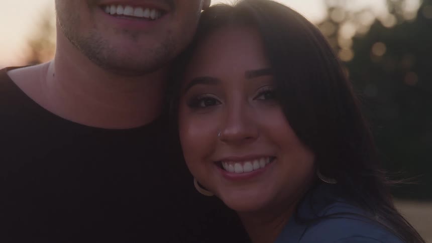 A young man with a slight beard and a young woman with long dark hair smile and stand close together looking at the camera