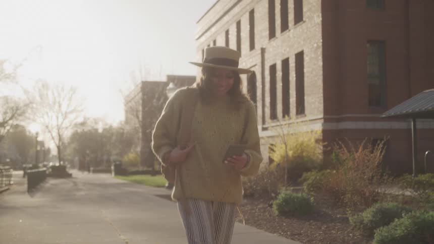 Woman wearing a hat and beige sweater walks on the sidewalk looking at her phone