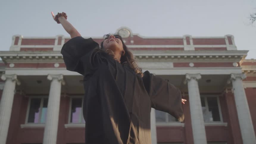 A woman in a blue graduation gown is happily tossing her cap in the air with a bright smile on her face with a brick building and columns visible in the background