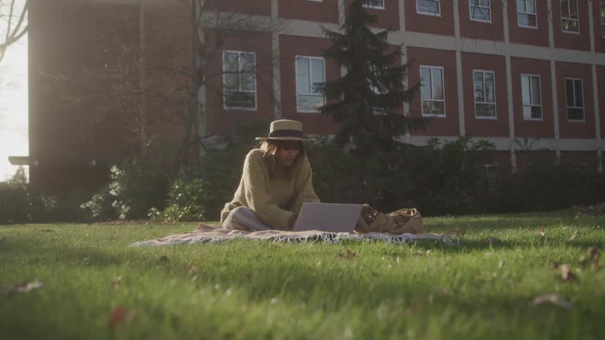 A woman wearing a sweater and a hat sits on a blanket on a grassy lawn in front of a brick building working on her laptop